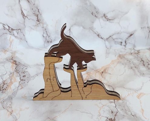 Layered wooden sculpture of a playful cat standing on a girl’s hand and foot, made from walnut, clear acrylic, and plywood, with a marble-effect white background