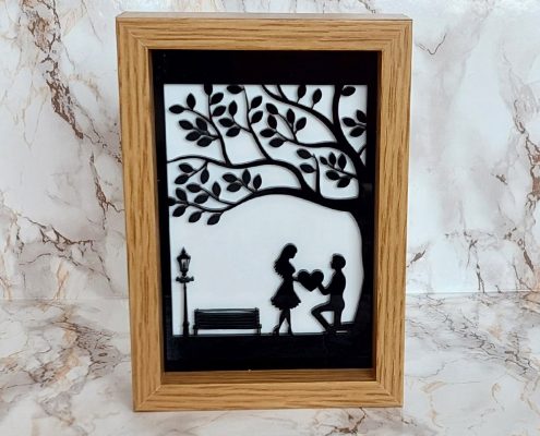 Romantic proposal silhouette art featuring black acrylic figures on white acrylic background in wooden frame - man proposing under tree